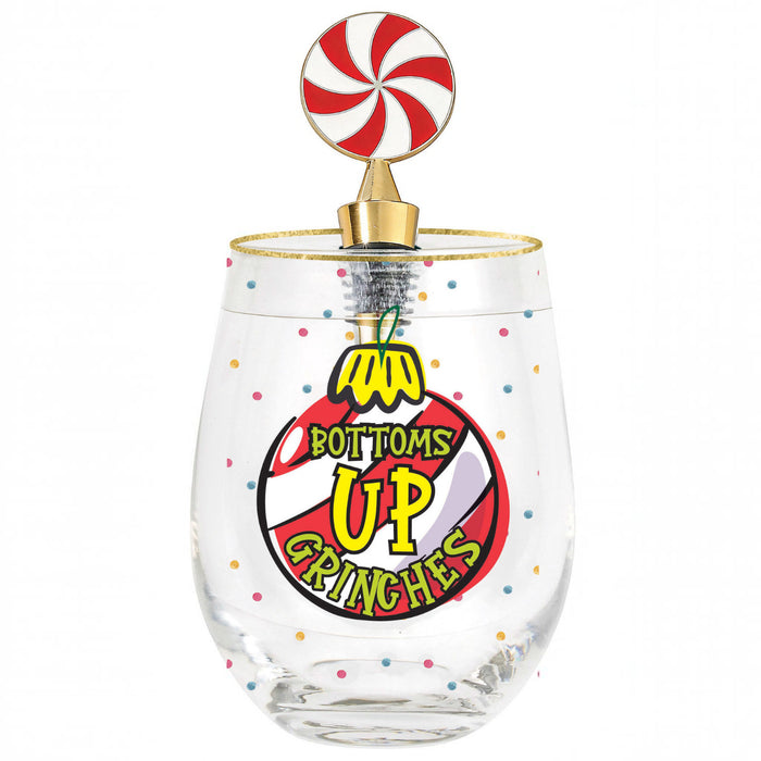 A clear stemless wine glass with a gold rim, decorated with colorful confetti dots and a large red, yellow, and black graphic that reads 'BOTTOMS UP GRINCHES' with a nutcracker illustration. It is topped with a gold-finished candy corkscrew featuring a red and white swirl lollipop on a white background.