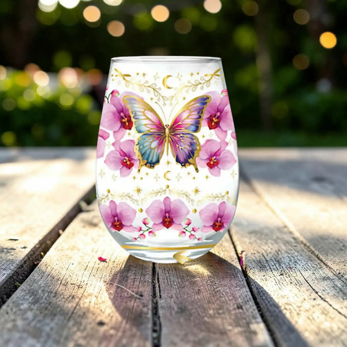 A clear stemless wine glass with a colorful printed design of a blue and purple butterfly, pink orchids, gold crescent moons, and stars, sitting on a weathered wooden table with a blurred green garden background.