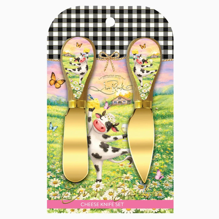 A cheese knife set in retail packaging, featuring two gold-colored cheese knives with decorative handles. The handles are decorated with a whimsical illustration of a black-and-white cow, butterflies, flowers, and a pastel sky.