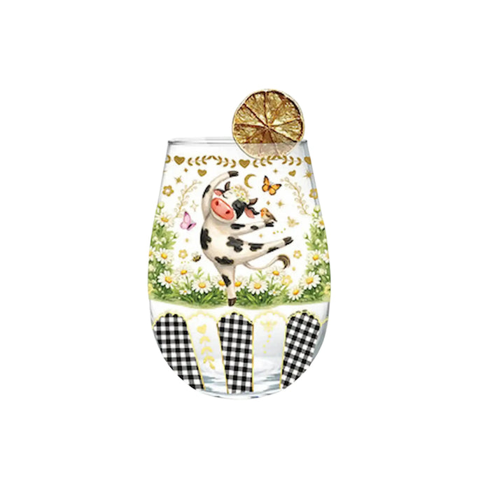 A stemless clear glass tumbler with a whimsical cow illustration, daisies, butterflies, and gingham pattern accents, shown on a plain white background with a dried lemon slice garnish.