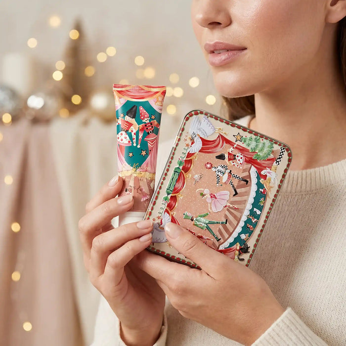 A woman holds a decorative tube and matching rectangular box with a whimsical illustrated design featuring a carousel, ballerinas, and festive colors. The background is softly blurred with warm bokeh lights.