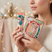A woman holds a decorative tube and matching rectangular box with a whimsical illustrated design featuring a carousel, ballerinas, and festive colors. The background is softly blurred with warm bokeh lights.