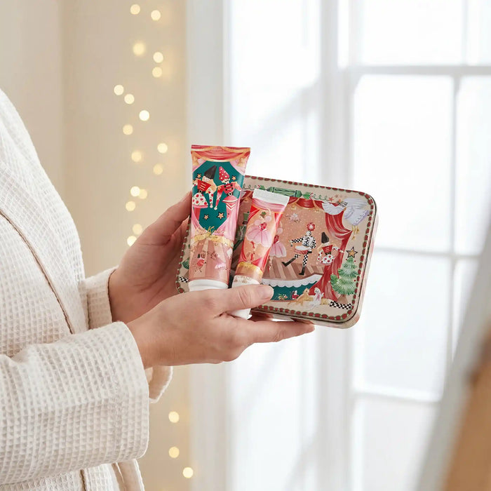A person in a cream waffle-knit robe holds a decorative tin containing two hand cream tubes with festive Christmas illustrations, set against a softly blurred background with warm string lights and a bright window.