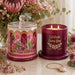 A decorative candle set displayed on a light marble surface with floral arrangements in the background. The set includes a clear glass jar candle with a lid and a burgundy glass candle vessel with gold text reading 'Celebrate Every Day' and a floral print.