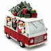A festive Christmas-themed toy or collectible vehicle featuring a red and white van with Santa Claus, dogs, a penguin, and a bear peeking out the windows, loaded with a small snowy Christmas tree and wrapped gifts on top, photographed on a white background.