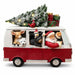 A festive red and white Christmas van figurine with Santa Claus, a deer, a polar bear, a penguin, and a small bird peeking out of the windows. A snow-dusted Christmas tree with warm lights and wrapped gifts sits on the roof, all on a clean white background.
