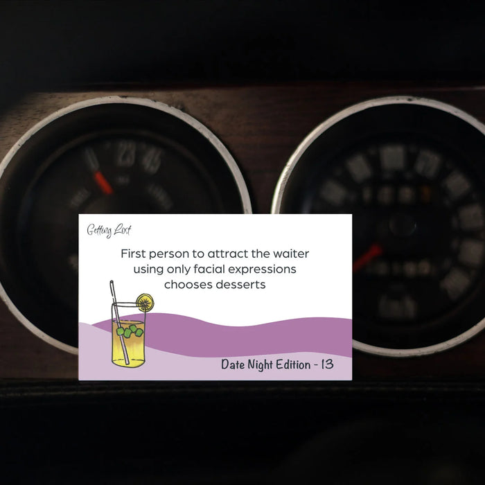 A rectangular novelty card or sign with printed text and a cocktail illustration is placed in front of two dark car dashboard gauges. The card reads 'Getting Loud' and 'First person to attract the waiter using only facial expressions chooses desserts' with 'Date Night Edition - 13' at the bottom.