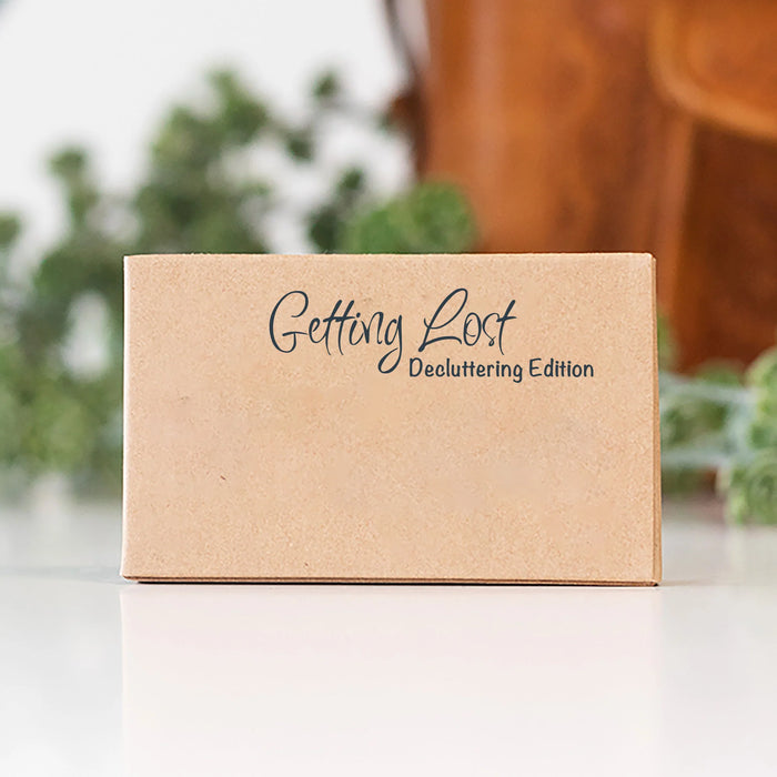 A small kraft paper box with the words 'Getting Lost' and 'Decluttering Edition' printed on the front, photographed on a white surface with a softly blurred indoor background.