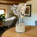 A clear glass reed diffuser bottle wrapped in beige burlap, with white laser-cut botanical reeds and a silver collar, displayed on a wooden table in a softly blurred home interior.