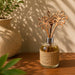 A glass reed diffuser bottle wrapped in burlap, filled with amber liquid, with three laser-cut wooden flower-shaped reeds. It sits on a wooden table in warm sunlight with a textured vase and plant in the background.