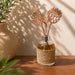 A glass reed diffuser bottle wrapped in natural jute, filled with amber liquid, and topped with two laser-cut wooden reeds shaped like leaves, displayed on a wooden table with warm sunlight and shadow patterns.