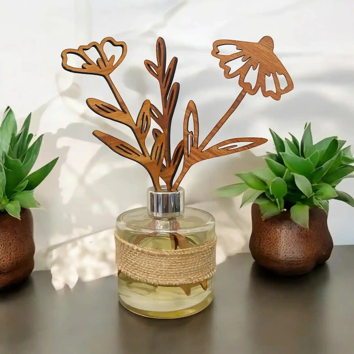 A glass reed diffuser bottle with a natural jute-wrapped neck and a laser-cut wooden flower diffuser top, styled on a tabletop between two potted succulents.