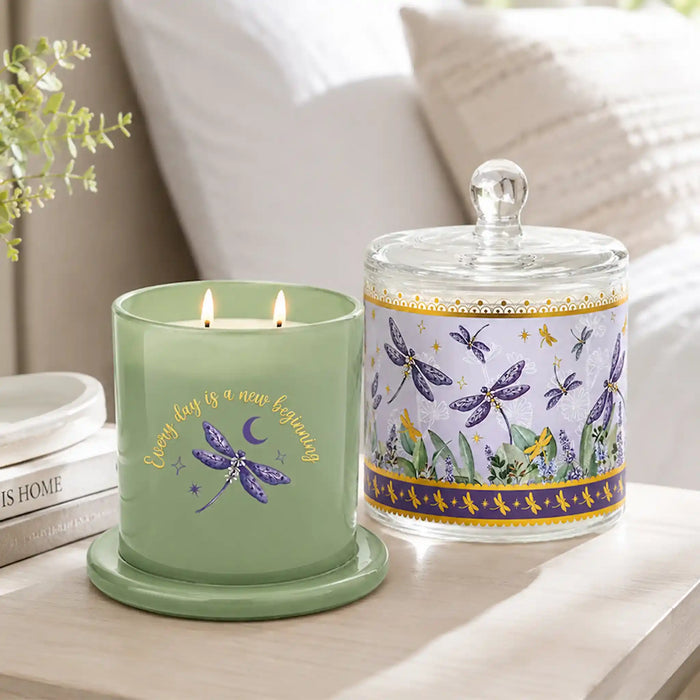 A lit sage green scented candle in a glass jar sits beside a matching glass jar with a lid, both decorated with purple dragonflies, yellow birds, and gold script text, set on a light wood table in a softly lit bedroom.