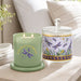 A lit sage green scented candle in a glass jar sits beside a matching glass jar with a lid, both decorated with purple dragonflies, yellow birds, and gold script text, set on a light wood table in a softly lit bedroom.