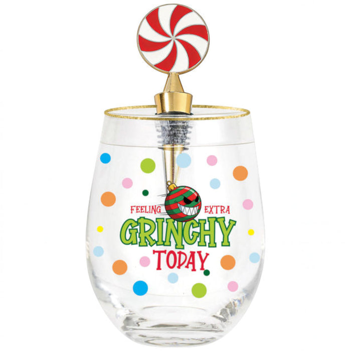 A clear stemless wine glass with a gold rim and a festive holiday-themed lollipop topper. The glass features colorful polka dots and the text 'FEELING GRINCHY EXTRA GRINCHY TODAY' in green and red, with a small ornament charm hanging from the topper.