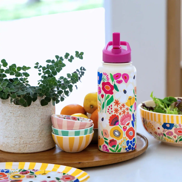 A white insulated water bottle with a colorful floral and bird pattern, topped with a bright pink lid, displayed on a wooden tray next to bowls, a plant, and fruit in a bright kitchen setting.