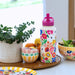 A white insulated water bottle with a colorful floral and bird pattern, topped with a bright pink lid, displayed on a wooden tray next to bowls, a plant, and fruit in a bright kitchen setting.
