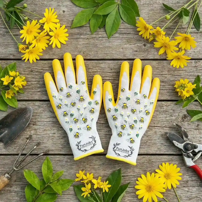 A pair of white and yellow gardening gloves with bee grip patterns, laid flat on a rustic wooden surface surrounded by gardening tools and yellow flowers. The gloves feature the FunJobs brand logo near the cuffs.
