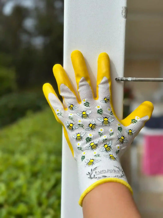 A close-up of a gardening glove with yellow palms and fingers, a white back, and a pattern of small yellow-and-black bees and white flowers, worn on a hand touching a white fence post outdoors.