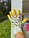 A close-up of a gardening glove with yellow palms and fingers, a white back, and a pattern of small yellow-and-black bees and white flowers, worn on a hand touching a white fence post outdoors.