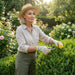 A woman wearing a straw hat, beige shirt, olive pants, and gardening gloves uses pruning shears to trim a green hedge in a sunlit garden.