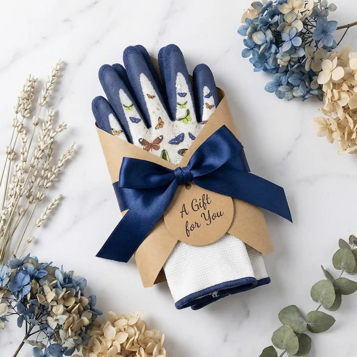 A gift set of gardening gloves and a folded white cloth wrapped in kraft paper, tied with a navy ribbon and a round tag that reads 'A Gift for You.' The gloves are navy blue with white back panels decorated with butterfly and leaf illustrations.