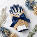 A gift set of gardening gloves and a folded white cloth wrapped in kraft paper, tied with a navy ribbon and a round tag that reads 'A Gift for You.' The gloves are navy blue with white back panels decorated with butterfly and leaf illustrations.