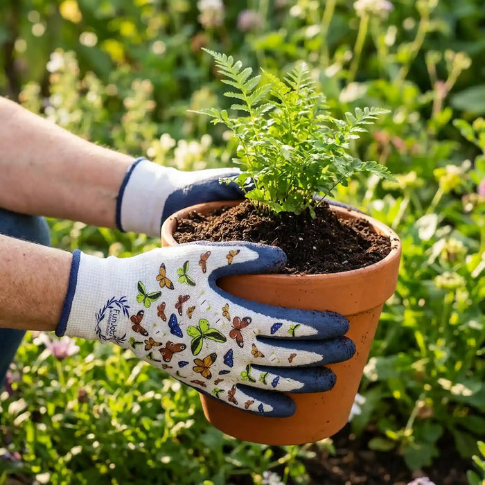 A person wearing white and navy gardening gloves decorated with colorful butterfly illustrations holds a small green plant in a terracotta pot, set against a blurred garden background.