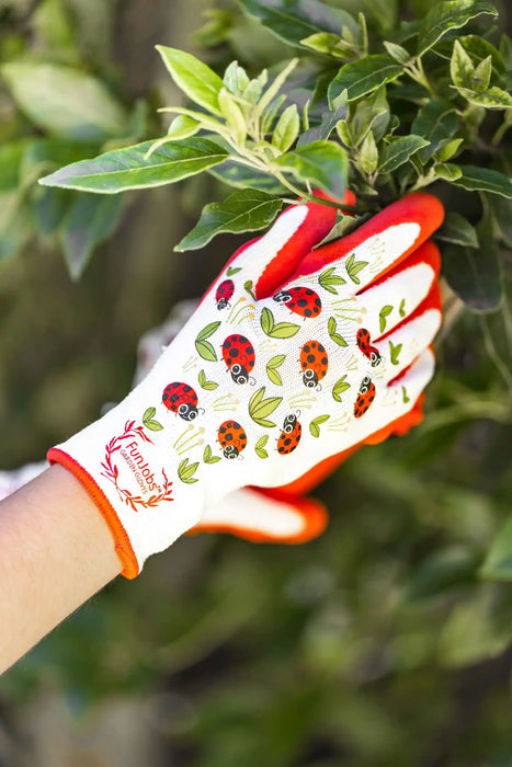 A gardening glove with a white back, red palm and fingertips, and a ladybug pattern, worn on a hand touching green leaves outdoors.