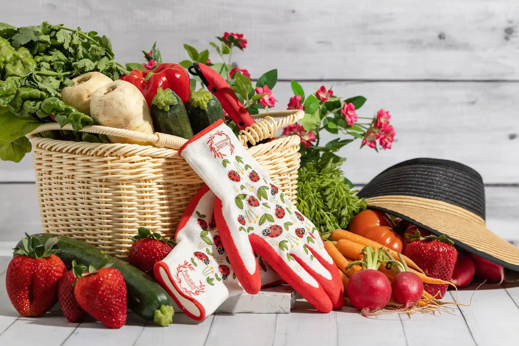 A pair of white and red oven mitts with strawberry and leaf patterns, styled in a rustic garden-themed scene with a wicker basket of vegetables, strawberries, radishes, a black straw hat, and a white wood plank background.