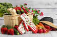 A pair of white and red oven mitts with strawberry and leaf patterns, styled in a rustic garden-themed scene with a wicker basket of vegetables, strawberries, radishes, a black straw hat, and a white wood plank background.