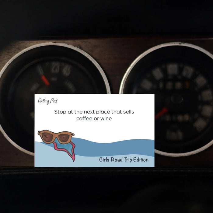 A rectangular printed card or sticker with a quote and illustration is placed in front of two dark vehicle dashboard gauges. The card reads 'Getting Lost' and 'Stop at the next place that sells coffee or wine' with 'Girls Road Trip Edition' at the bottom.