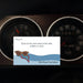 A rectangular printed card or sticker with a quote and illustration is placed in front of two dark vehicle dashboard gauges. The card reads 'Getting Lost' and 'Stop at the next place that sells coffee or wine' with 'Girls Road Trip Edition' at the bottom.