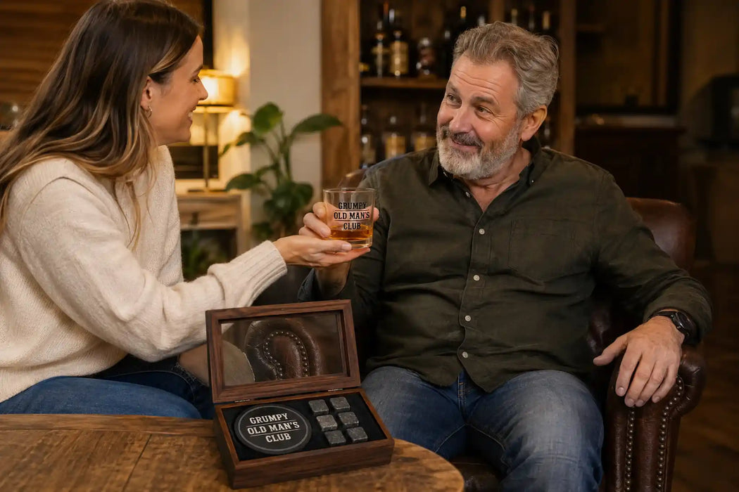 A man and a woman sit in a warmly lit bar or lounge setting, holding glasses of amber liquor. In the foreground, an open wooden box labeled 'Grumpy Old Man's Club' sits on a table.