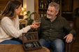 A man and a woman sit in a warmly lit bar or lounge setting, holding glasses of amber liquor. In the foreground, an open wooden box labeled 'Grumpy Old Man's Club' sits on a table.