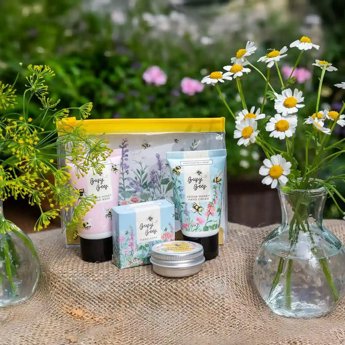 A set of beeswax body care products from 'Beeswax Soap' arranged on a burlap table outdoors with daisies in glass vases. The set includes a pink soap bar in a clear pouch with a yellow border, a blue hand cream tube, a small blue box, a small round tin, and a larger blue soap bar.