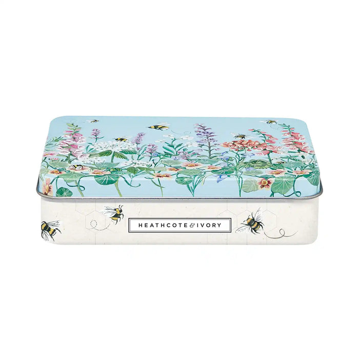 A rectangular metal tin with rounded corners, featuring a botanical illustration on the lid with pink flowers, green leaves, and bees over a light blue background. The front of the tin is white with a black label reading 'HEATHCOTE & IVORY'.