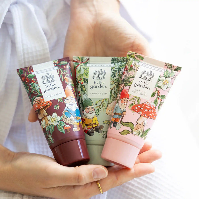 Three tubes of 'In the Garden' hand and foot creams held in a person's hands against a white ribbed knit background. The tubes feature whimsical garden gnome illustrations and are shown in dark burgundy, sage green, and light pink colors.
