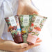 Three tubes of 'In the Garden' hand and foot creams held in a person's hands against a white ribbed knit background. The tubes feature whimsical garden gnome illustrations and are shown in dark burgundy, sage green, and light pink colors.