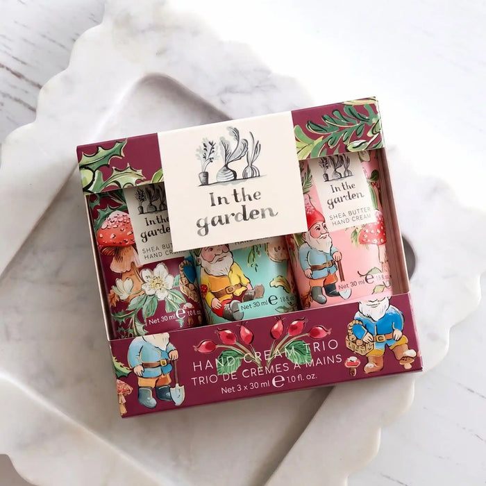 A boxed set of three hand creams in a decorative gift box labeled 'In the garden,' placed on a white marble tray over a light wood surface. The packaging features colorful garden-themed illustrations and text for hand cream trio.