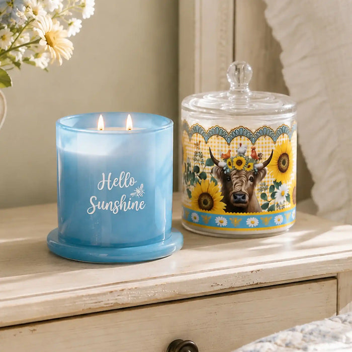 A lit blue candle in a glass holder sits on a light wood dresser beside a decorative glass jar with a cow and sunflower illustration. The scene is styled in a bedroom or living space with soft natural light and flowers in the background.