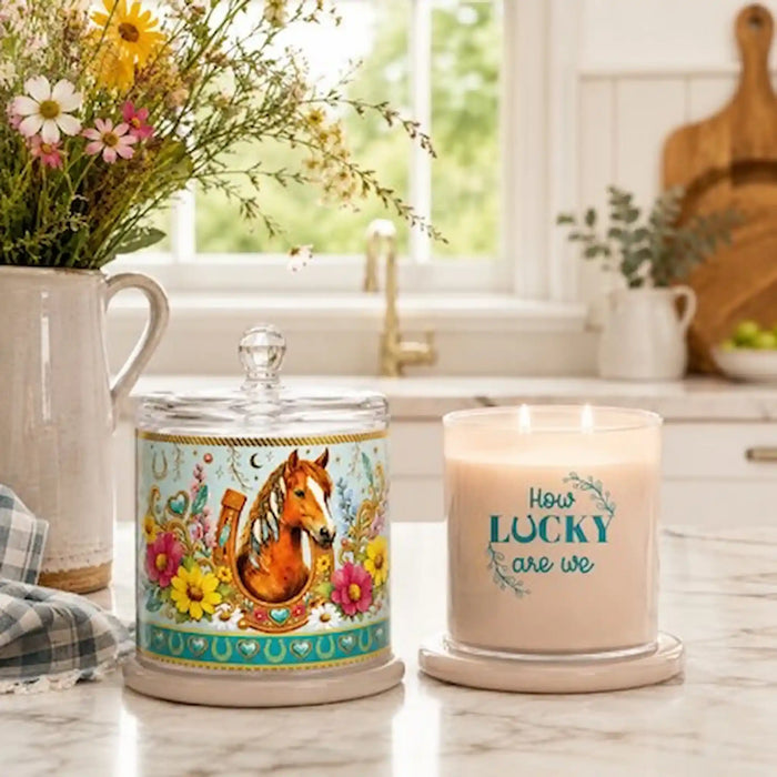Two decorative candles on a kitchen countertop: a lidded glass jar candle with a colorful horse illustration and a lit cream candle in a glass holder that reads “How LUCKY are we.” A vase of flowers and a blurred kitchen background are visible.