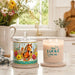 Two decorative candles on a kitchen countertop: a lidded glass jar candle with a colorful horse illustration and a lit cream candle in a glass holder that reads “How LUCKY are we.” A vase of flowers and a blurred kitchen background are visible.