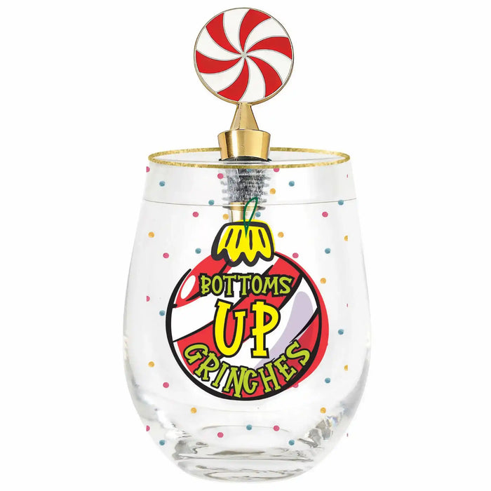 A clear stemless wine glass with a gold rim and a decorative top featuring a red and white swirl candy lollipop on a gold post. The front of the glass is printed with a colorful graphic that reads 'BOTTOMS UP GRINCHES' with a yellow penguin and red, black, and white circular design, accented with small multicolored confetti dots.