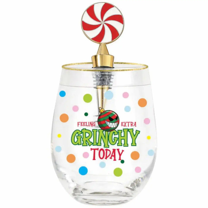 A clear stemless wine glass with a gold rim and a festive holiday-themed lollipop topper. The glass features colorful polka dots and the text 'FEELING GRINCHY EXTRA GRINCHY TODAY' in green and red, with a small ornament charm hanging from the topper.