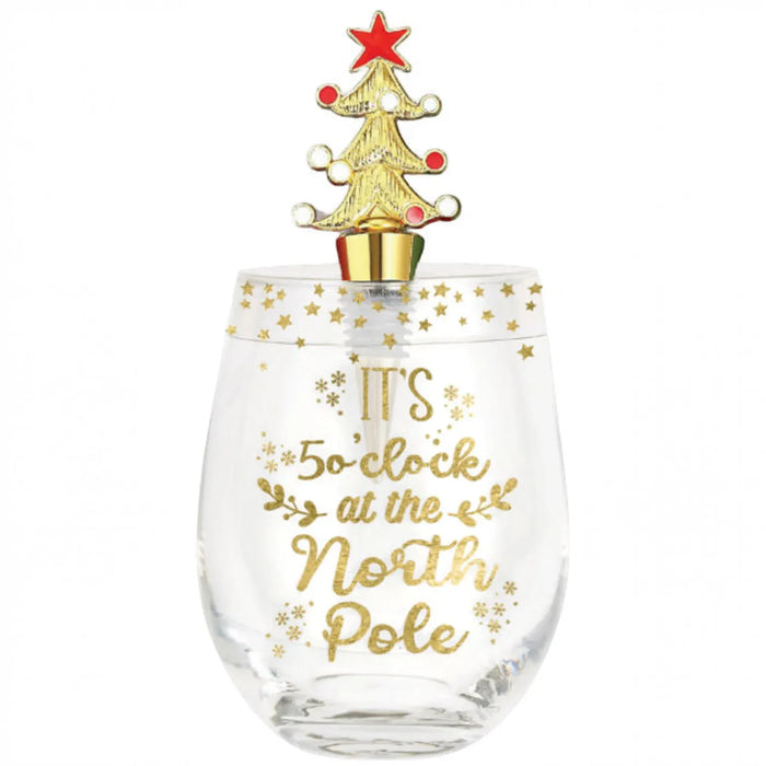 A clear stemless wine glass with gold foil text reading 'IT'S 5:00 at the North Pole' and gold star/snowflake accents, topped with a gold Christmas tree ornament featuring a red star.