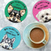Four round dog-themed coasters arranged on a light marble-like surface, with a cup of coffee on a saucer in the foreground. The coasters feature illustrated dog portraits and the phrase 'ALL YOU NEED IS LOVE - AND A [dog breed]'.