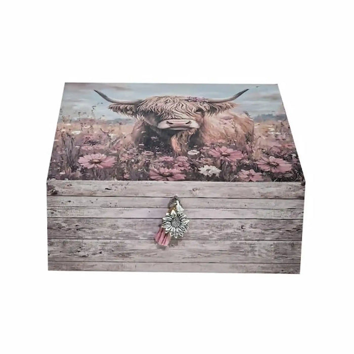 A rectangular wooden storage box with a rustic weathered gray wood-grain finish on the front and lid, featuring a printed illustration of a highland cow in pink wildflowers. A small decorative tassel ornament hangs from the front clasp against a plain white background.