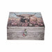 A rectangular wooden storage box with a rustic weathered gray wood-grain finish on the front and lid, featuring a printed illustration of a highland cow in pink wildflowers. A small decorative tassel ornament hangs from the front clasp against a plain white background.