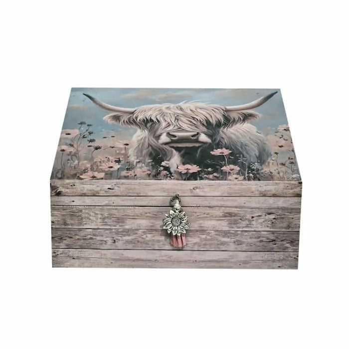 A rectangular wooden storage box with a rustic weathered wood grain finish, featuring a printed illustration of a shaggy Highland cow in a field of pink flowers on the lid. The front has a decorative silver-toned metal clasp with a red tassel, set against a plain white background.